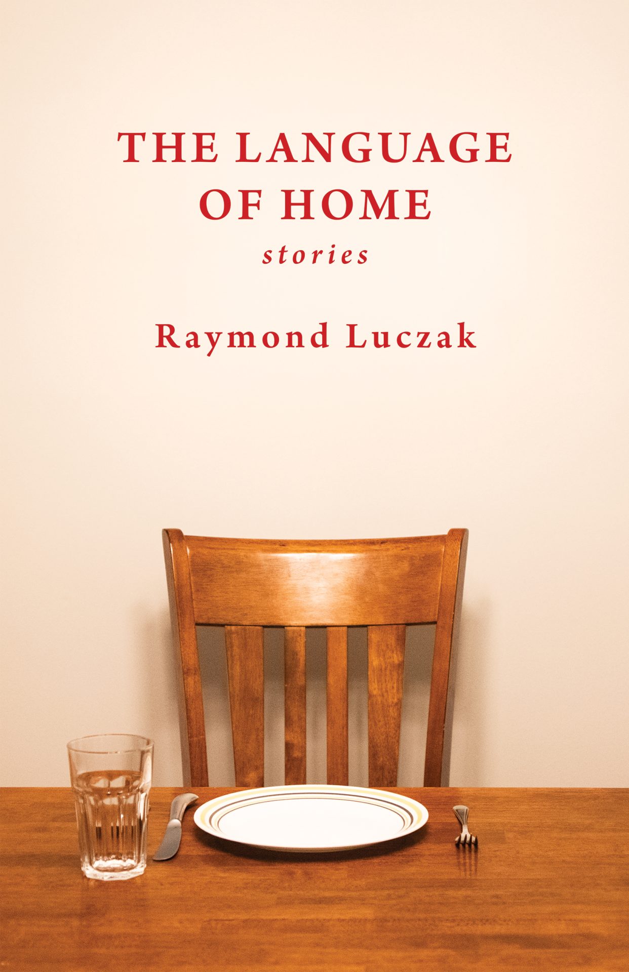 Against an off-white wall lit brightly from above, a heavily varnished brown wooden chair is set before a table that’s equally heavily varnished. Before the chair is a white plate with two slender circles painted right near the plate’s edge. On either side of the plate is a silver fork and knife; on the left side of the plate is an empty glass. Higher up on the wall above the chair is the following text in dark red: THE LANGUAGE OF HOME; stories; Raymond Luczak.