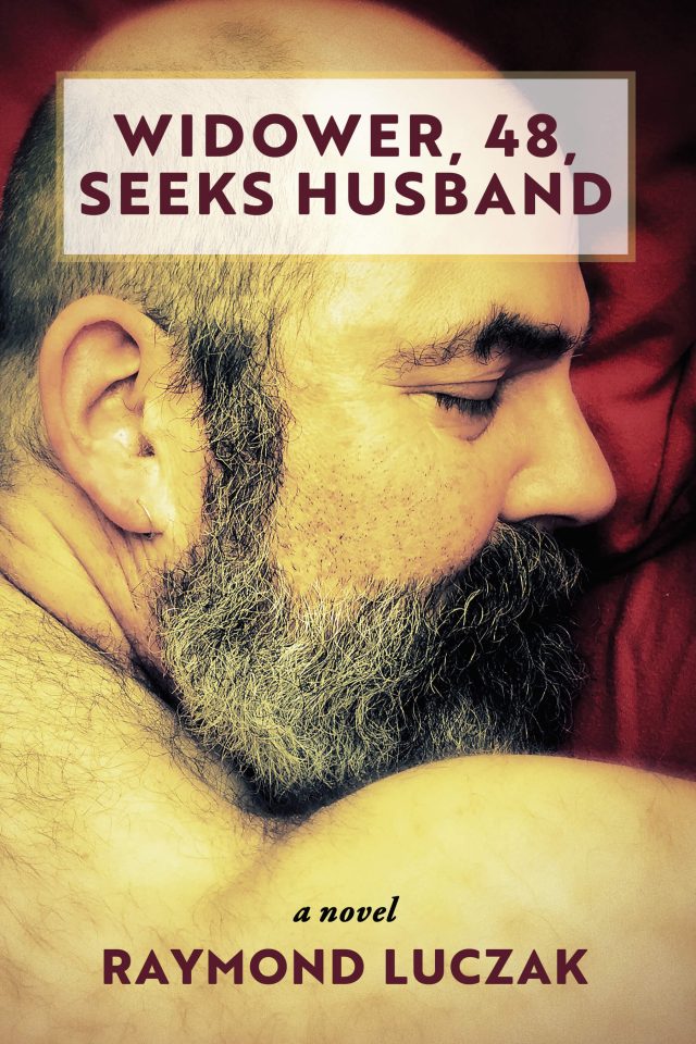 In a soft golden light, a balding bearded middle-aged man closes his eyes while he cuddles a pillow sideways in red flannel. Across the top of his head is a early opaque white box with the title WIDOWER, 48, SEEKS HUSBAND in a dark mauve color. Across the man's shoulder near the bottom of the cover is the text "a novel" in black italics and the author's name Raymond Luczak in a dark mauve color.